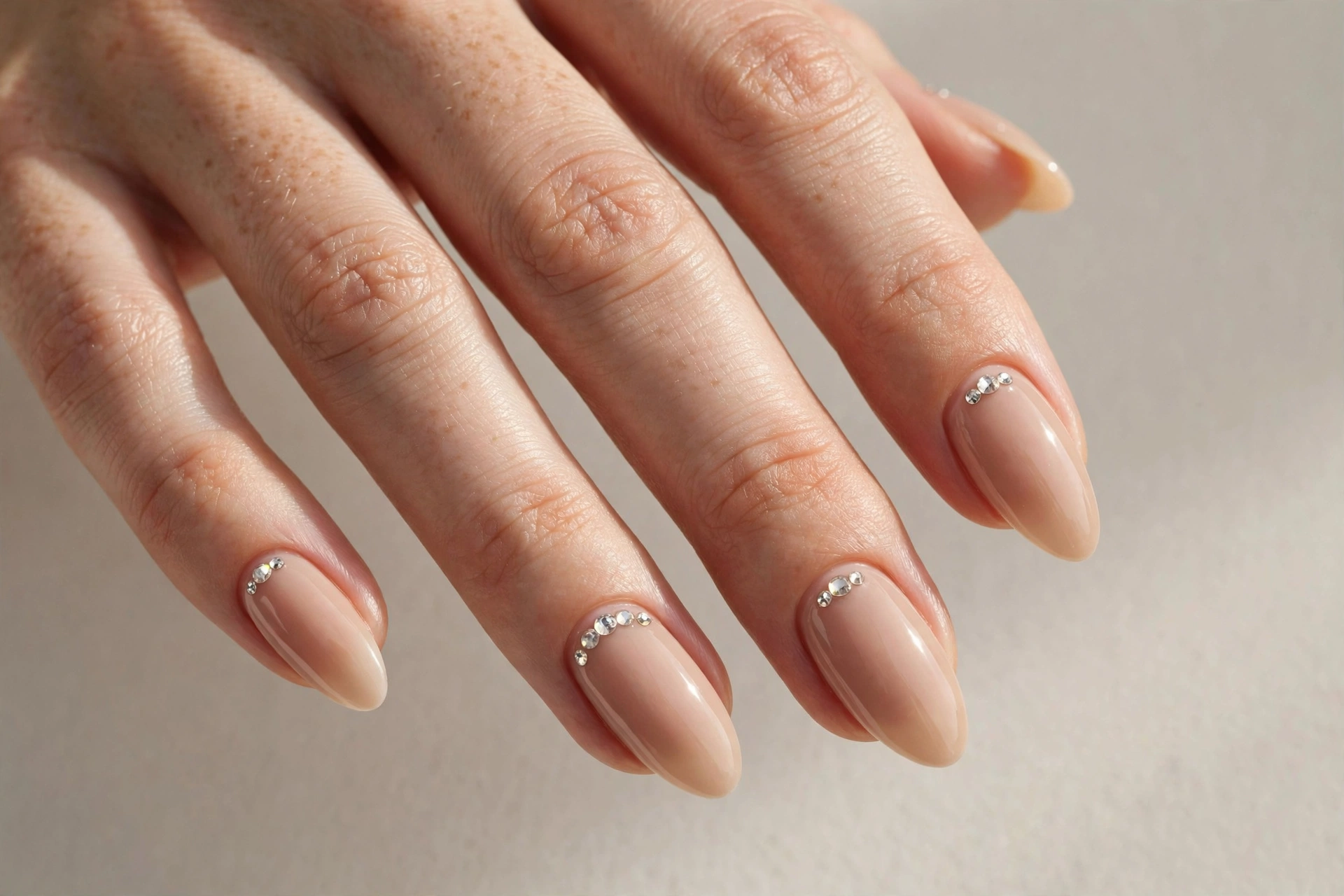 Close-up of a woman's hand with nude almond-shaped acrylic nails decorated with small crystal rhinestones