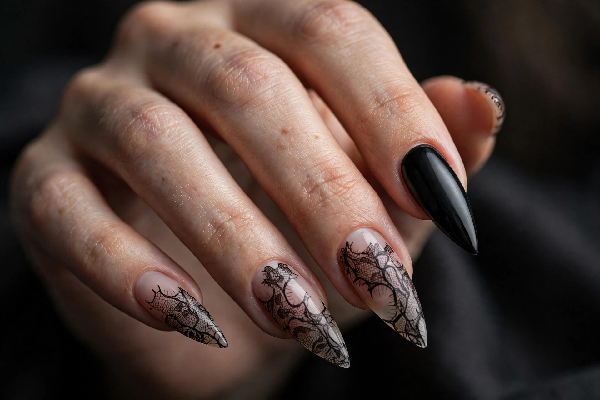 Sheer nude stiletto nails with delicate black lace pattern overlay and one solid black accent nail