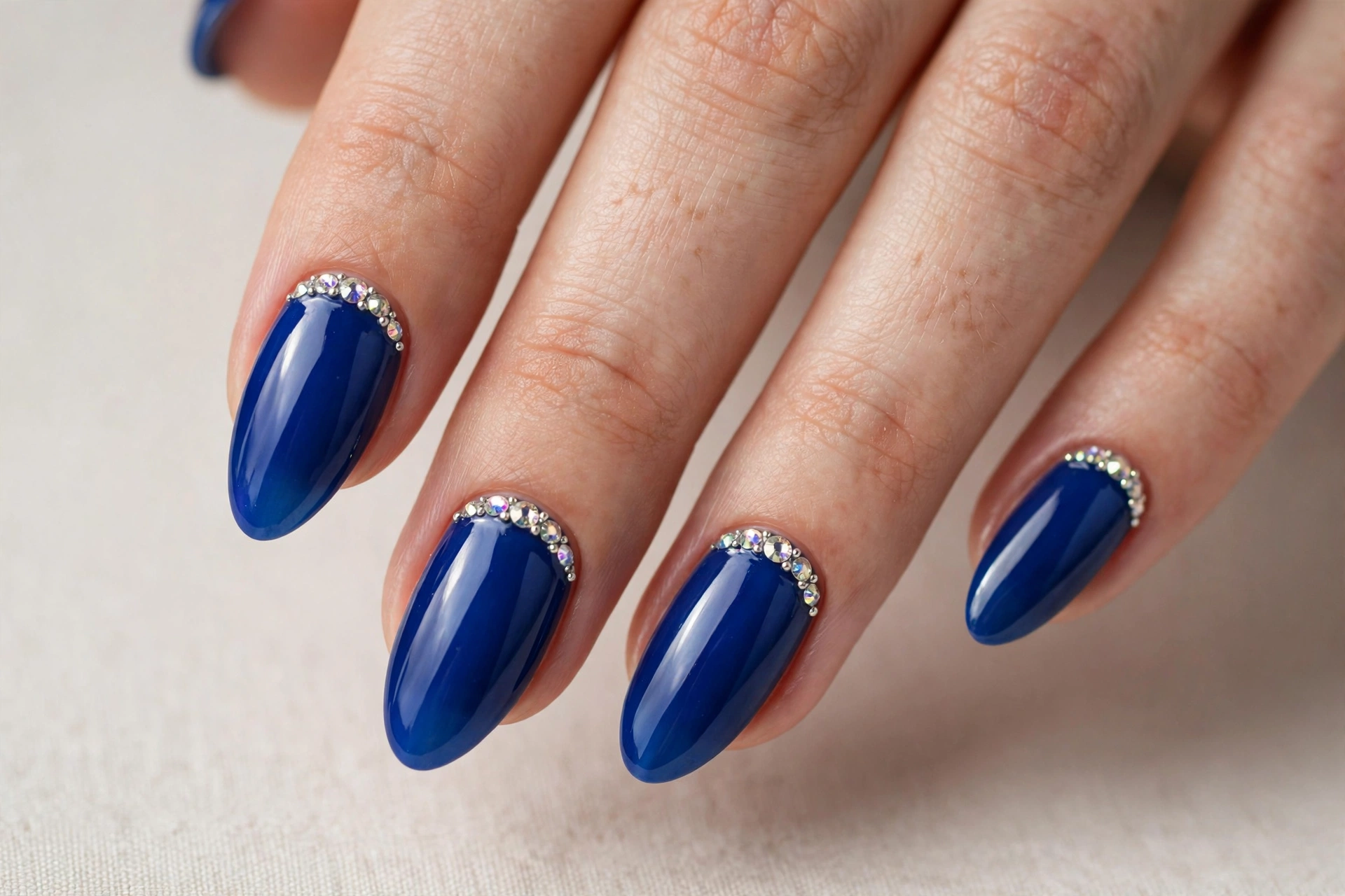 Sapphire blue manicure with sparkling rhinestone accents at the cuticles on almond nails