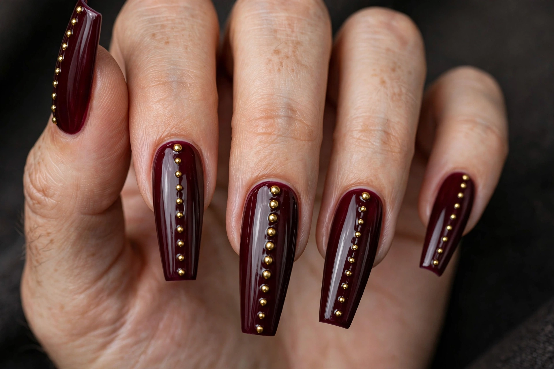 Long coffin nails in glossy deep burgundy polish with small gold stud rivets in a line down the center
