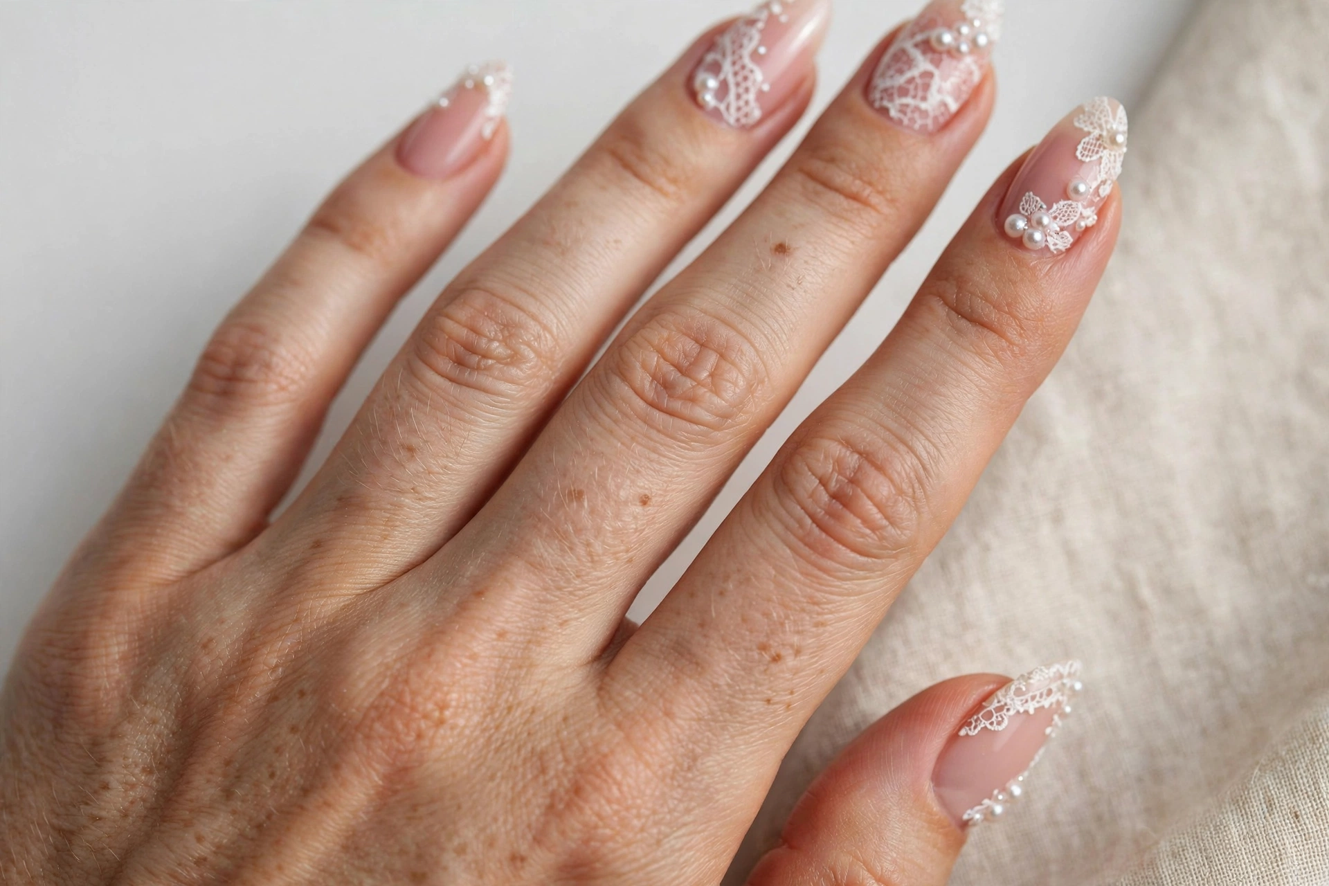 White lace nail art with tiny pearl accents on soft pink nude almond nails