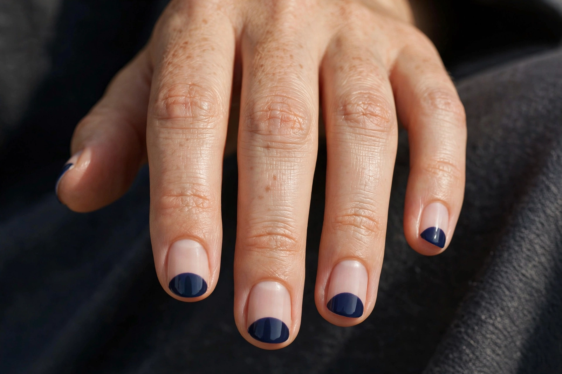 Short round nails with nude base and painted half-moon shapes at the cuticle in deep navy